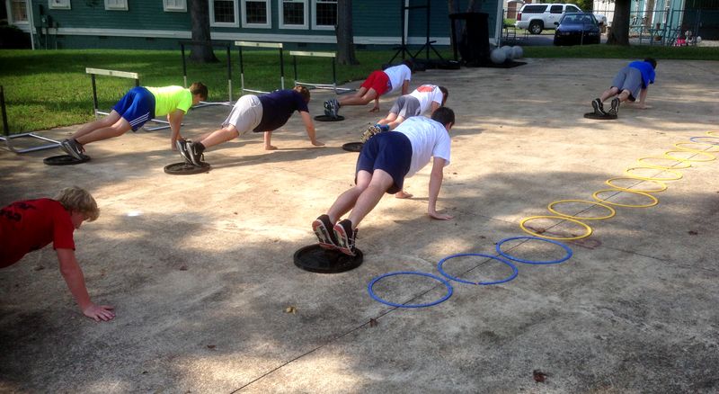 Youth athletes doing outdoor plank drills beside agility rings and hurdles